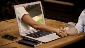 Auf dem Foto schütteln sich zwei Menschen die Hand. Eine Person sitzt vor dem Laptop und die andere Person streckt den Arm aus dem Laptop.